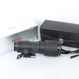 Puede incluir: Un objetivo teleobjetivo Sigma negro con una funda de transporte de cuero negro. El objetivo tiene un anillo plateado alrededor del frontal y un anillo de zoom plateado. La funda tiene una hebilla plateada y una correa negra.