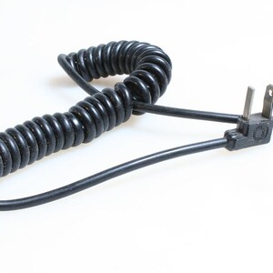 Pode incluir: Cabo de alimentação preto enrolado em espiral com uma ficha de dois pinos e um conector de três pinos. O cabo é enrolado em espiral e a ficha é do tipo padrão.
