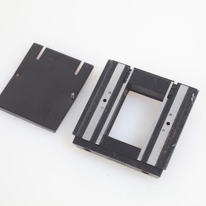 May include: A black rectangular component with a textured surface and two vertical slots, next to a larger black and silver frame. The frame has a rectangular opening and silver stripes. The components appear to be made of plastic.