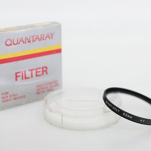 May include: A black camera lens filter in a clear plastic case with a white box that says "QUANTARAY FILTER" on it. The filter is labelled "QUANTARAY 62mm +1 JAPAN".