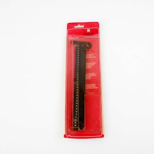 Op de afbeelding: Een zwarte bindkam in een rode en transparante plastic verpakking. De kam is lang en heeft een reeks kleine gaatjes langs de rug. De verpakking heeft tekst. Het product staat op een witte achtergrond.