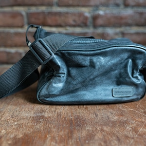 May include: A black leather shoulder bag with a zipper closure and a brand label that reads "QUANDARY".