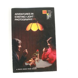 May include: A vintage book cover titled "Adventures in Existing-Light Photography" featuring a couple at a table, illuminated by a warm-toned lamp. The cover has a black background with the Kodak logo and a price tag.