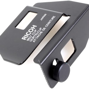 May include: Black metal belt clip with a silver inscription that reads "RICOH BELT CLIP GÜRTELCLIP ATTACHE DE CEINTURE JAPAN".
