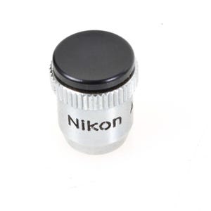 Puede incluir: Un objetivo de cámara Nikon con una tapa negra. El cuerpo del objetivo es plateado con el nombre de la marca "Nikon" impreso. El objetivo tiene un agarre estriado y texturizado para facilitar su manejo. El fondo es blanco.