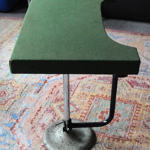 May include: A vintage green-topped adjustable table with a curved edge. The table has a chrome-colored, adjustable central pole and a round, silver-colored base. A black, curved support arm extends from the pole to the table's edge.
