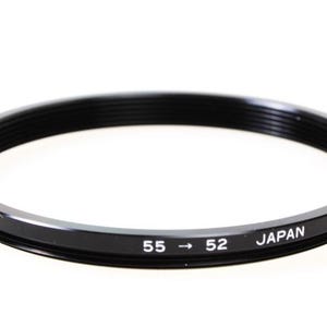 May include: Black step-down ring with the text "55 -> 52 JAPAN" printed in white. The ring is circular with a threaded interior and a smooth, glossy exterior. The ring is designed for photography.