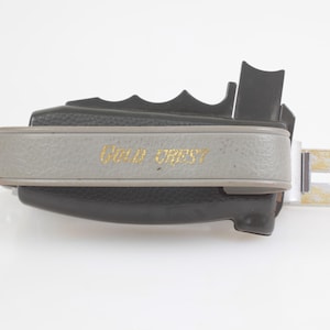Puede incluir: Una cámara vintage en blanco y negro con las palabras "Gold Crest" en letras doradas en la banda gris. La cámara tiene forma rectangular con una pieza metálica plateada que se extiende desde el lateral.