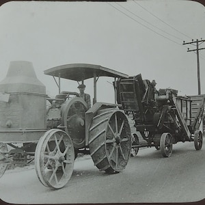 May include: A vintage black and white photograph of a large agricultural tractor with prominent metal spoked wheels and a canopy, pulling a mechanical harvesting implement along a road, with a person visible in the driver seat area.
