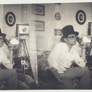 May include: A black and white photo of a person wearing a top hat and a white shirt, sitting in a room with a camera on a tripod. The person is looking at the camera and holding a hand to their chin.