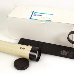 May include: A beige Spiratone telephoto lens with black accents, a lens cap, and a black mounting bracket. The lens is next to a white box with a blue and white label. The lens is approximately 25 cm long.