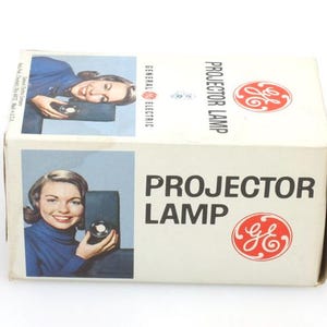 May include: Vintage GE Projector Lamp box. The box is white with the words "PROJECTOR LAMP" in black. The GE logo is in red. The box features images of a woman holding a projector. The box is on a white background.