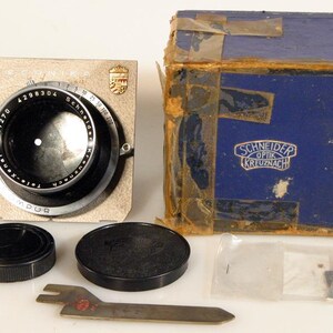 May include: Vintage camera lens with a black and silver design, alongside a blue box with the text "Schneider Kreuznach". Includes lens caps, a tool, and small accessories. The lens has the text "Technika" and "Tele-Xenar".