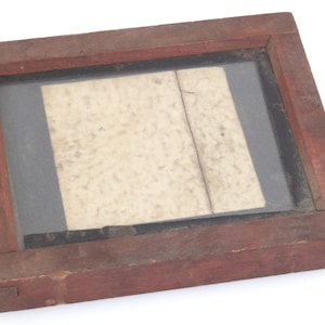 May include: A rectangular wooden frame with a glass pane, enclosing a piece of aged paper. The frame is stained a reddish-brown colour, and the paper has a faded, textured appearance. The paper is divided by a vertical line.