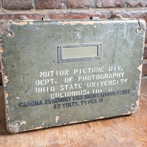 Puede incluir: Una caja de metal verde con una etiqueta que dice "MOTION PICTURE DIV. DEPT. OF PHOTOGRAPHY OHIO STATE UNIVERSITY COLUMBUS, 10, 0 CAMERA ASSEMBLY GUN SIGHT AIMING POINT 12 VOLTS, TYPES M".