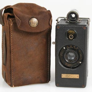 May include: Vintage black camera with a lens, viewfinder, and a small brass plate. A brown leather case with a button closure is next to the camera. The camera has a circular lens and a dial.