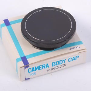 May include: A black camera body cap with a ridged edge sits atop a box. The box is off-white with blue and purple stripes and text that reads "CAMERA BODY CAP FOR MINOLTA" and "JAPAN".