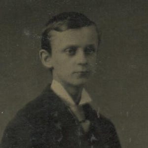 May include: A vintage black and white portrait of a young person. The subject is wearing a dark suit jacket over a white collared shirt. The image has a slightly faded appearance, typical of older photographs.