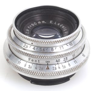 May include: A vintage camera lens with a silver metal body and black accents. The lens has multiple rings with numbers and text, including "f=50 mm" and "Feet". The lens is designed for photography.
