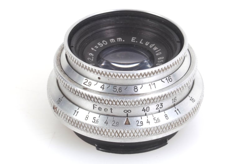 May include: A vintage camera lens with a silver metal body and black accents. The lens has multiple rings with numbers and text, including "f=50 mm" and "Feet". The lens is designed for photography.
