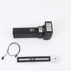 Puede incluir: Un flash negro con una cabeza de flash blanca y un cable negro. El flash está unido a un soporte de metal plateado.