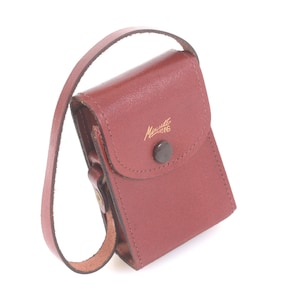 May include: A burgundy leather case with a strap, featuring a flap closure secured by a dark button. The case has a small gold inscription on the front. The strap is attached to the top of the case.