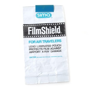 May include: White FilmShield pouch with blue clip, designed for air travelers. The pouch protects film from airport X-ray damage. The text "FilmShield" is prominently displayed.