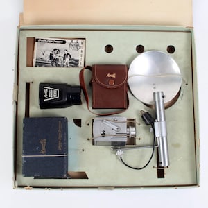 Puede incluir: Kit de cámara vintage en una caja. El kit incluye una cámara negra, una funda de cuero marrón, un reflector de flash plateado y un pequeño folleto de instrucciones. La caja es de color verde claro con recortes para cada artículo.