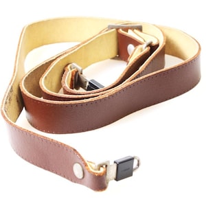 May include: A brown leather camera strap with a light tan interior. The strap has metal hardware and a button closure. The leather appears to be stitched along the edges. The strap is designed to be worn around the neck or shoulder.