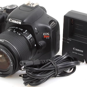 May include: A black Canon EOS Rebel T2i digital SLR camera with a Canon EF-S 18-55mm lens attached. The camera is on a white surface with a black Canon battery charger.