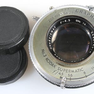 May include: A vintage camera lens with the text "GRAPHIC" and "KODAK SUPERMATIC" visible. The lens is silver and black, with a series of numbers and markings around the edge. Two black lens caps are stacked on the left.