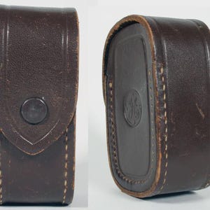 May include: A dark brown leather case with a rounded rectangular shape and visible stitching along the edges. The front features a pointed flap with a circular snap button closure, while the side displays a circular embossed logo with the letters GE.