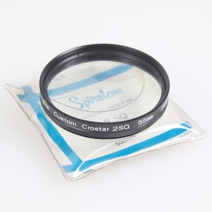 Op de afbeelding: Een zwarte Spiratone Custom Crostar 2SQ 55mm cameralensfilter. Het filter is cirkelvormig met een helder glazen midden en is verpakt in een doorzichtige plastic hoes met blauwe accenten. De tekst "Spiratone" is zichtbaar.