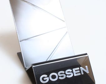 Puede incluir: Un soporte de plástico negro con una superficie reflectante plateada y la palabra "Gossen" impresa en letras plateadas.