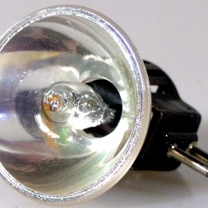 May include: A close-up of a silver projector lamp with a small bulb inside a reflective housing. The lamp has a black connector with two silver pins extending from it. The lamp is designed to project light.
