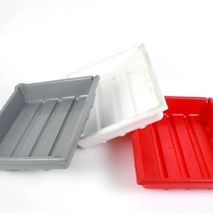 May include: Three rectangular plastic trays stacked at an angle, featuring a gray tray on the left, a white tray in the center, and a red tray on the right. Each tray has a smooth finish with three parallel ridges running along the bottom surface.