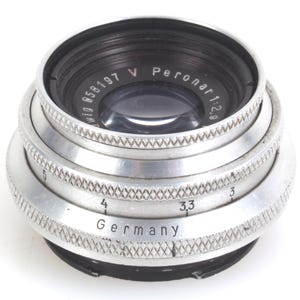 May include: Close-up of a vintage camera lens with a silver metal body and black accents. The lens has the text "Peronar 1:2.9" and "Germany" engraved on it. The lens has a series of textured rings.