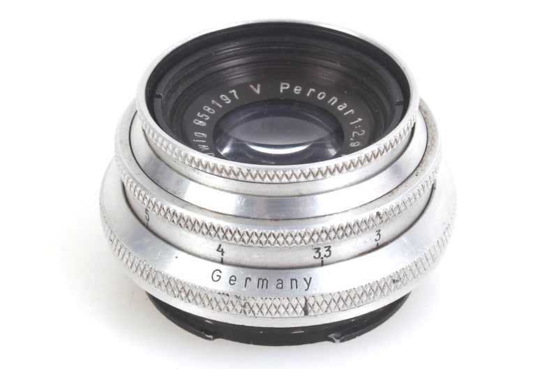May include: Close-up of a vintage camera lens with a silver metal body and black accents. The lens has the text "Peronar 1:2.9" and "Germany" engraved on it. The lens has a series of textured rings.
