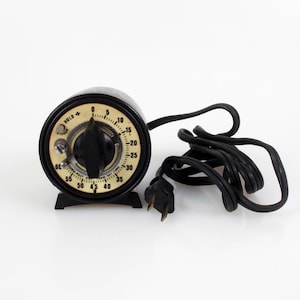 May include: A black electric kitchen timer with a round cream-colored face featuring black numerals from 0 to 60. The device has a central black rotating knob, a small metal switch, and a black power cord with a two-prong plug attached to the side.