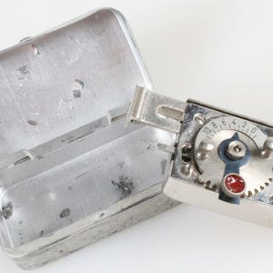May include: A silver metal timer with a round dial and a red button. The timer is in a silver metal case with a hinged lid.