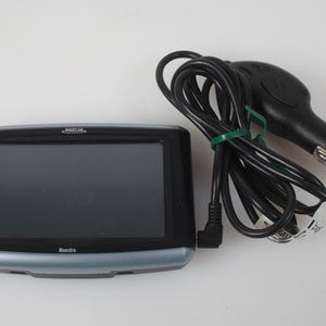 May include: A black and silver Magellan Maestro GPS navigation device with a rectangular screen and a black power cable featuring a car cigarette lighter adapter plug. The device has the brand name Magellan and the model name Maestro printed on it.