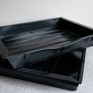 May include: Two black plastic trays stacked on top of each other. The trays have drainage holes and ridges on the bottom.