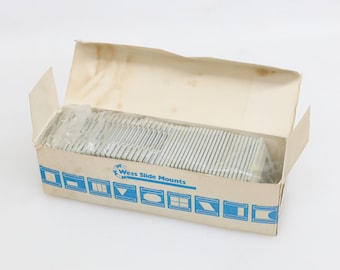 Può includere: Una scatola bianca con le parole "Wess Slide Mounts" stampate sul davanti. La scatola è aperta e contiene una pila di supporti per diapositive in plastica trasparente.