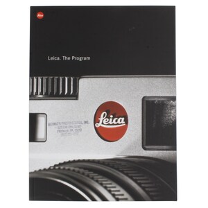 May include: A close-up of a Leica camera, featuring a silver body with black accents. The Leica logo is prominently displayed in a red circle. The text "Leica. The Program" is visible on the top of the camera.