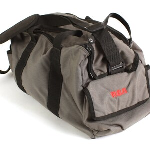 May include: A grey canvas duffel bag with black straps and a small grey pocket with the word "RCA" in red lettering.