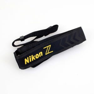 May include: Black Nikon Z camera strap with yellow lettering. The strap has a chevron pattern and a smaller adjustable strap. The Nikon Z logo is prominently displayed in yellow.