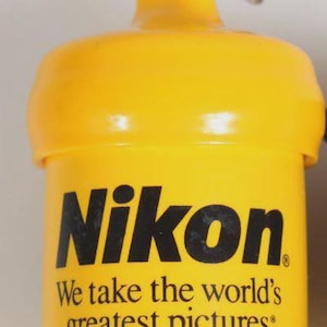 May include: A bright yellow, acorn-shaped plastic keychain featuring the black Nikon logo and the text, "We take the world's greatest pictures: Yours." A translucent white plastic loop and a silver metal split ring are attached to the top.