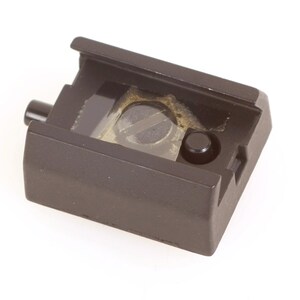 May include: A dark brown camera accessory, rectangular in shape, with a sliding top and a small, cylindrical button. Inside, a clear, rectangular component with a screw is visible. The item is on a white background.