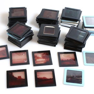 May include: A collection of vintage slide film negatives in black plastic frames. The slides are stacked in piles, some with a light blue frame.