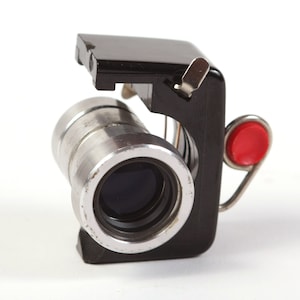 Puede incluir: Una cámara pequeña, negra y plateada con un botón rojo. La cámara está diseñada para ser utilizada con una tira de película.